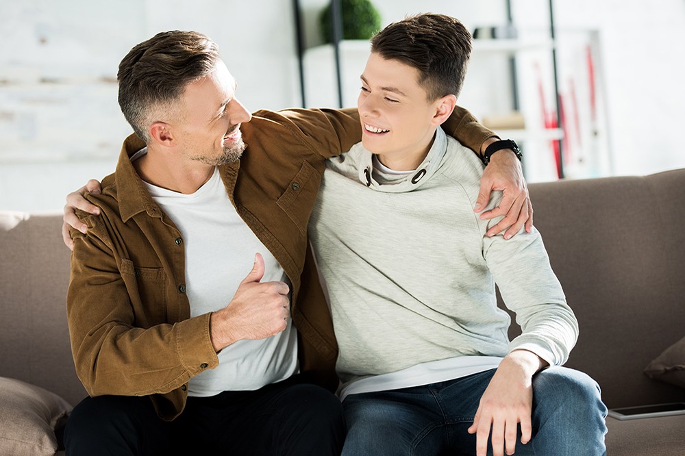 smiling father and teen son hugging on sofa at home and looking at each other