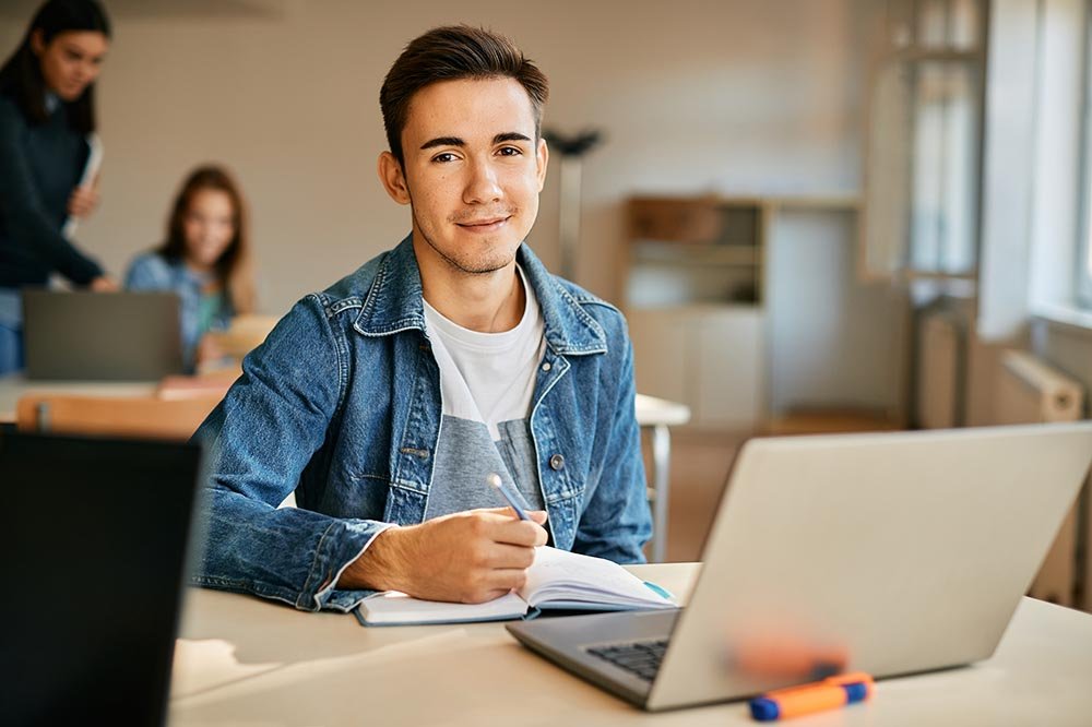 Smiling high school student e-learning on laptop and writing notes in the classroom while looking at camera.